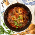 A pot of slow cooker osso buco, featuring braised meat with bone in a tomato-based sauce and garnished with fresh parsley, sits on a table with herbs, wooden utensils, and a blue-patterned plate nearby.