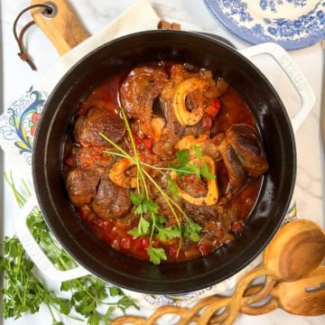 A pot of slow cooker osso buco, featuring braised meat with bone in a tomato-based sauce and garnished with fresh parsley, sits on a table with herbs, wooden utensils, and a blue-patterned plate nearby.