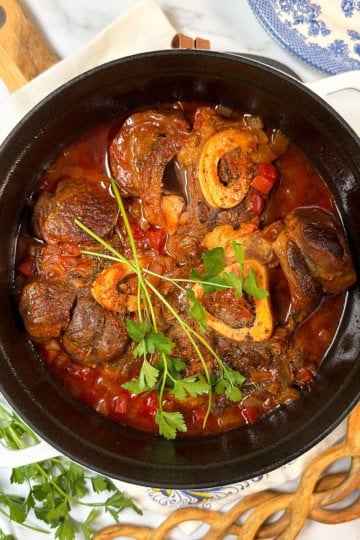 A pot of slow cooker osso buco, featuring braised meat with bone in a tomato-based sauce and garnished with fresh parsley, sits on a table with herbs, wooden utensils, and a blue-patterned plate nearby.