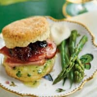 A biscuit sandwich with ham, a green speckled patty, and cherry jam elegantly presented on a decorative plate-perfect for Kentucky Derby recipes. Served with asparagus, green beans, and a dollop of sauce on the side. A glass of amber liquid gleams in the background.