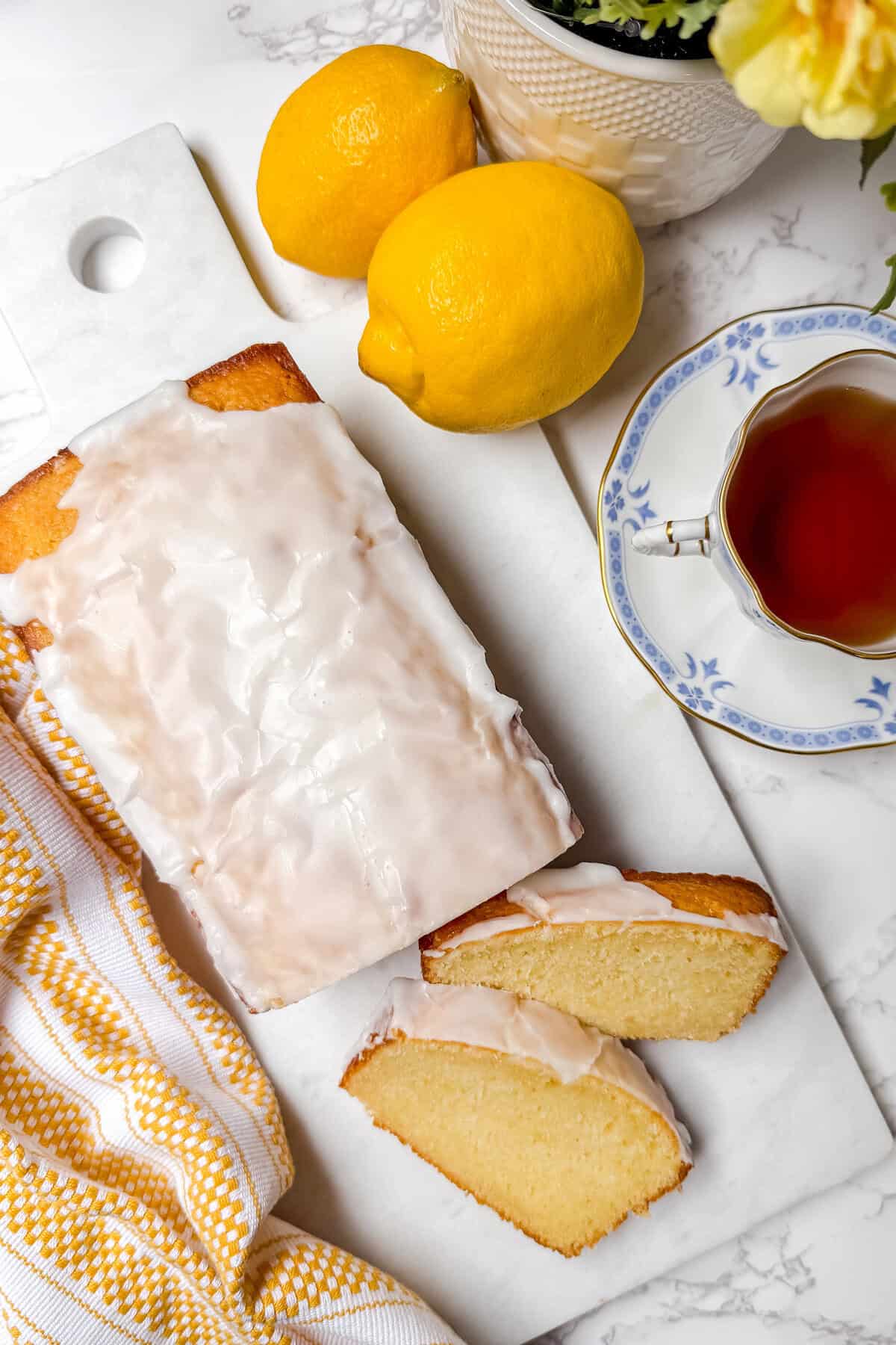 A lemon loaf cake with white icing sits on a marble board, flanked by two lemons, while two slices reveal its moist interior. Accompanied by a floral-patterned teacup filled with tea and a cheerful yellow-striped cloth, this scene is perfect for savoring the moment.
