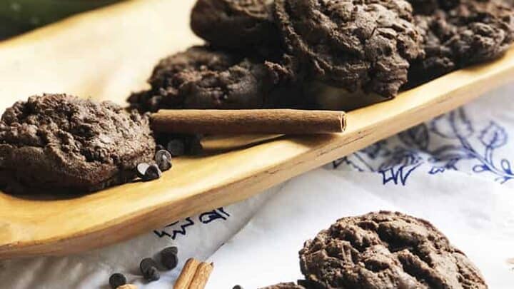 A wooden tray filled with Mexican chocolate cookies sits on a table. Some cookies are placed on a white surface with cinnamon sticks and scattered chocolate chips, enhancing their rich flavor. In the background, a pot holds additional cinnamon sticks.