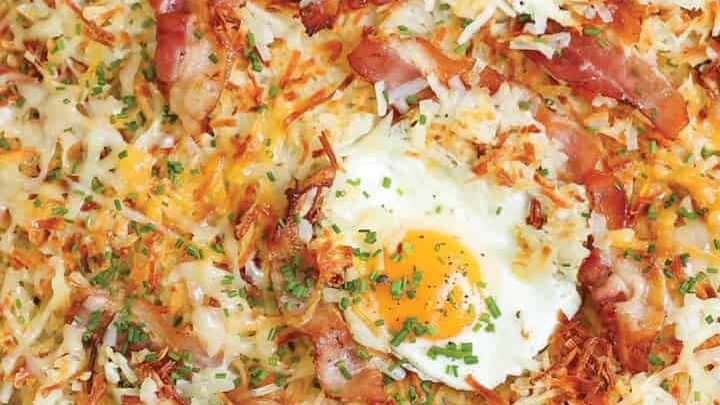 A sheet pan breakfast featuring crispy hash browns, topped with slices of bacon, melted cheese, and baked eggs. Finished off with a sprinkle of chopped chives for a pop of green color.
