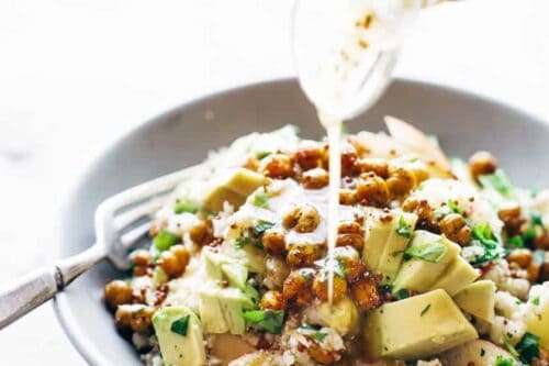 A hand drizzles dressing from a glass container onto a bowl of healthy salad featuring roasted chickpeas, diced avocado, brown rice, and greens. A fork rests on the edge of the gray bowl.