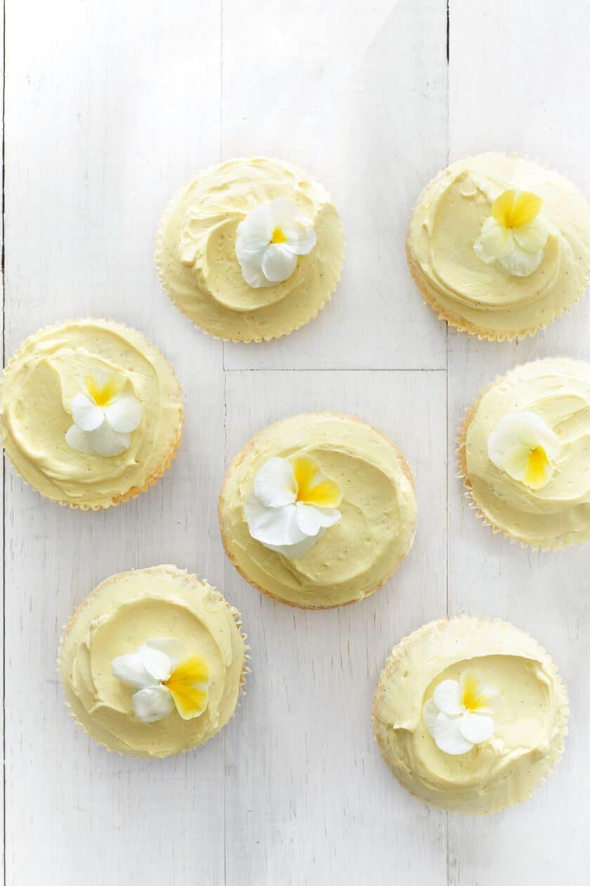 Seven vanilla cupcakes with creamy yellow frosting and delicate white and yellow flowers grace a white wooden surface. Perfect for spring brunch, these desserts are viewed from above, highlighting their exquisite decorative details.