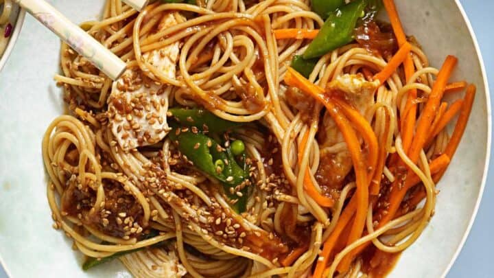 A bowl of noodles with shredded chicken, sliced carrots, snap peas, sesame seeds, and brown sauce—this healthy 20 minute dinner is served with a pair of chopsticks resting on the side.