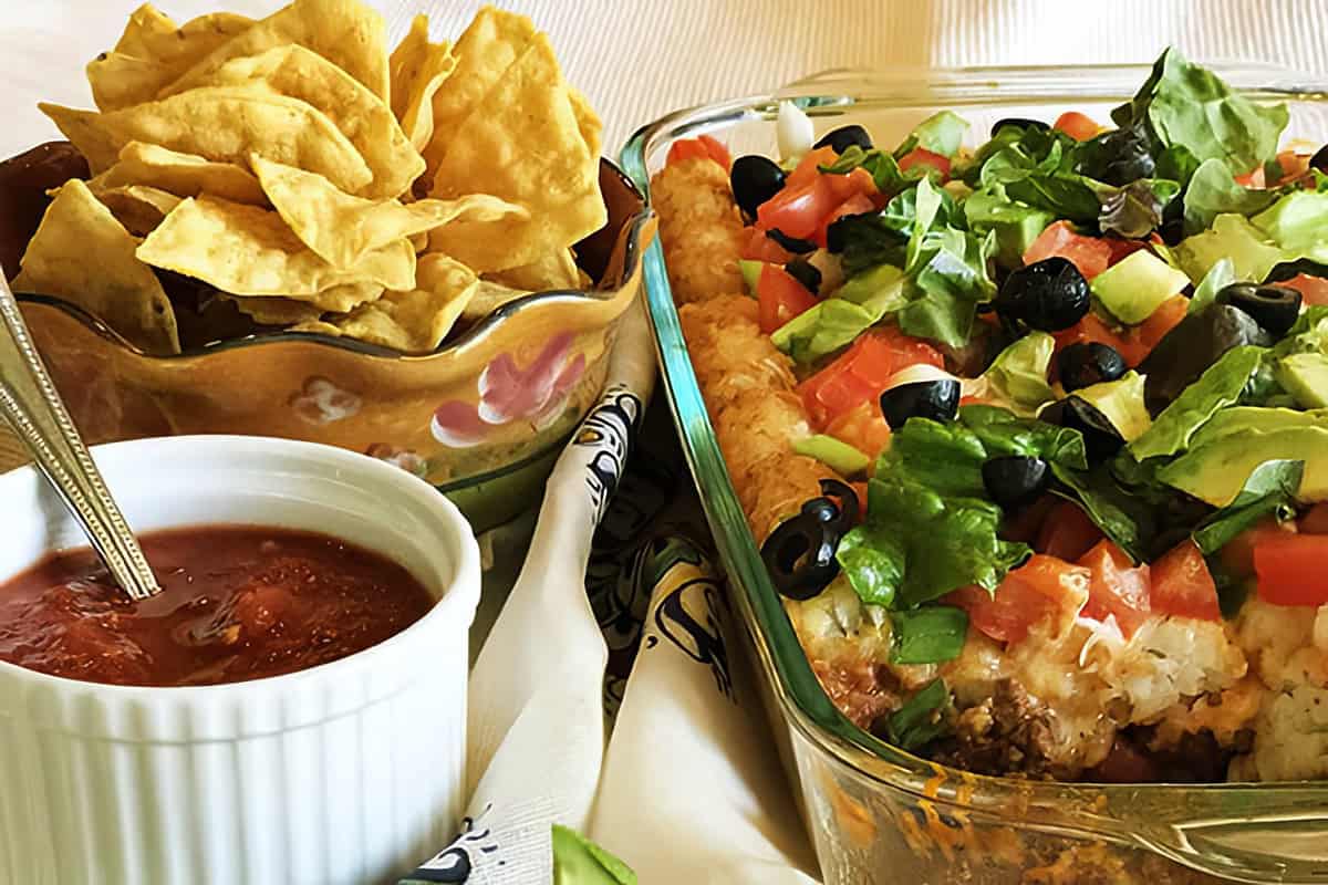 A Mexican-inspired dish of layered taco tater tot casserole topped with lettuce, tomatoes, black olives, and cheese fills a glass baking dish. Beside it sit a bowl of salsa and tortilla chips on a patterned tablecloth, echoing the hearty appeal of a Taco Tater Tot Casserole.