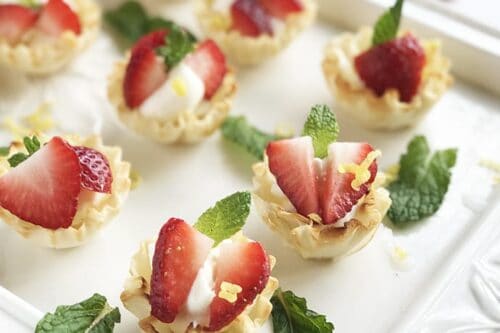 Mini strawberry cheesecakes elegantly presented as pastry cups filled with luscious cream, crowned with fresh strawberry slices and mint leaves, arranged on a white tray.