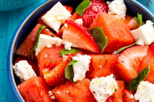A bowl of salad featuring watermelon cubes, sliced strawberries, feta cheese, and fresh basil leaves makes a refreshing choice for summer side dishes, all served atop a blue wooden table.
