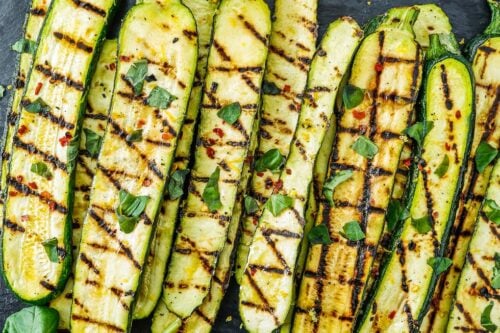 Grilled zucchini halves arranged on a dark slate board, garnished with herbs, served with lemon wedges and a small bowl of red pepper flakes—perfect for light Summer Side Dishes.