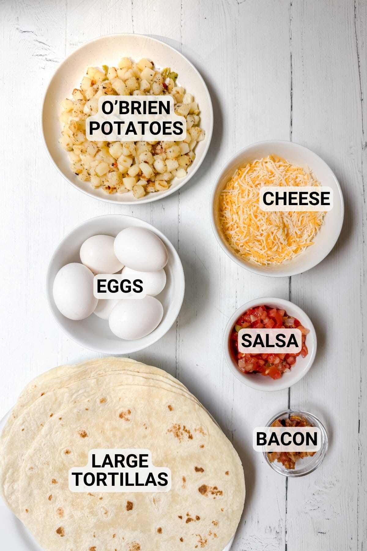 Breakfast burrito ingredients on white table including tortillas, eggs, potatoes, cheese, salsa, and bacon.