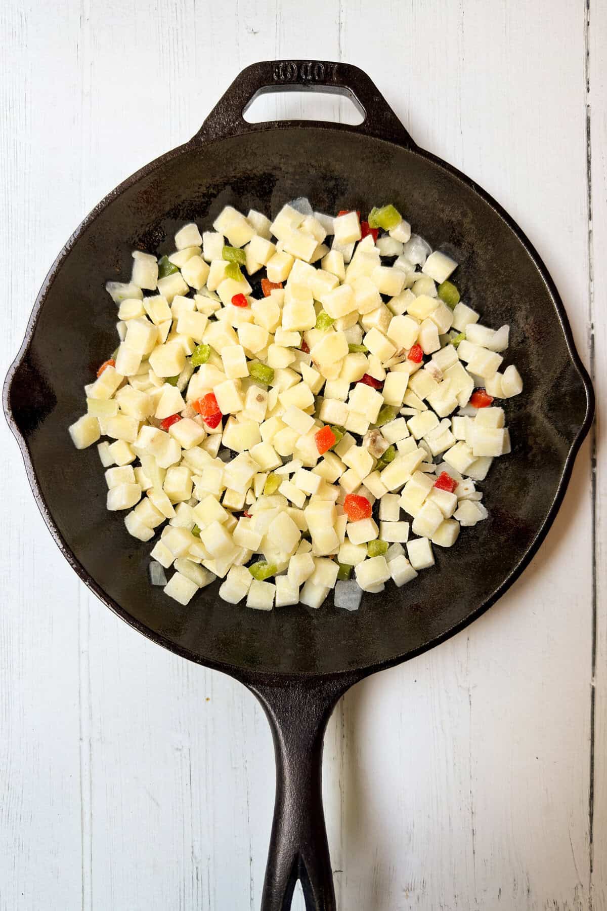 A cast iron skillet filled with diced potatoes and small pieces of red and green bell peppers, set on a white wooden surface-perfect fillings for hearty breakfast burritos.