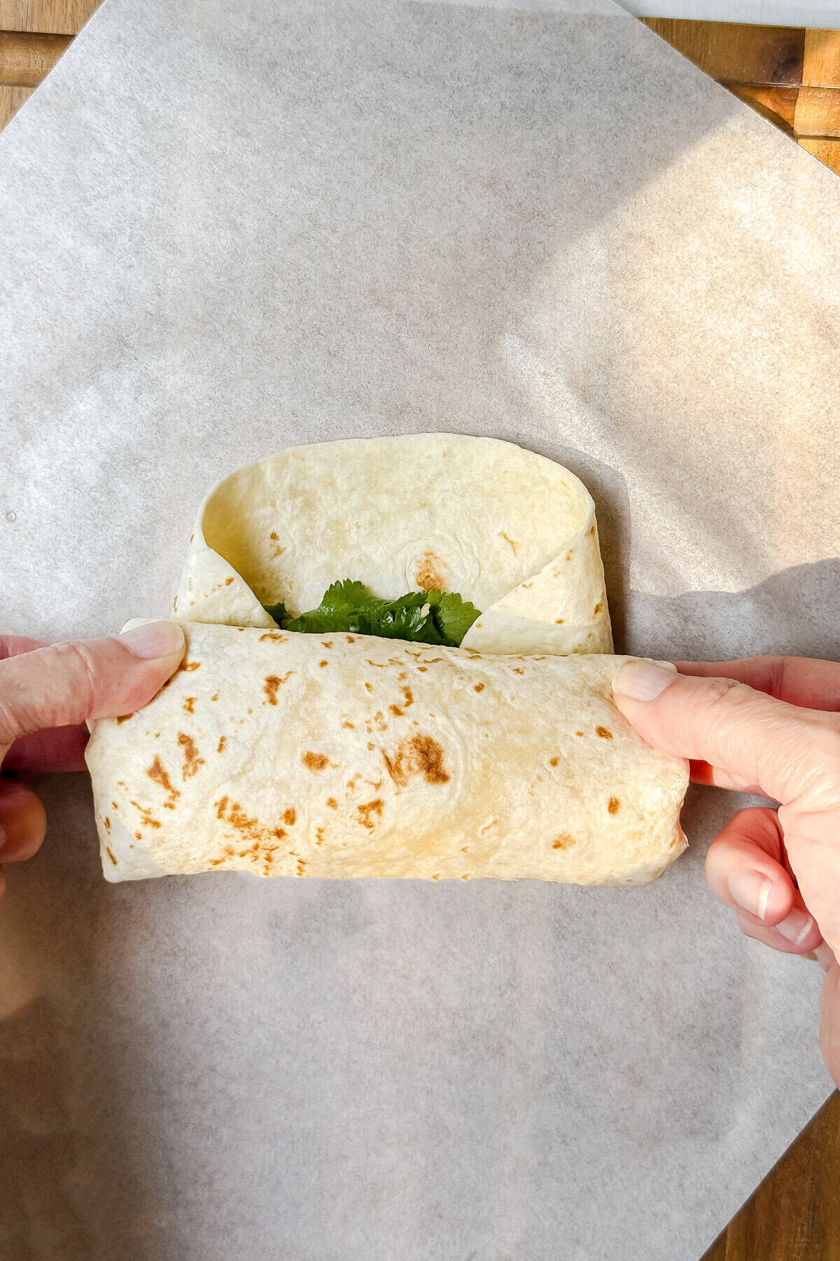 Hands folding a tortilla around breakfast burrito filling on parchment paper.