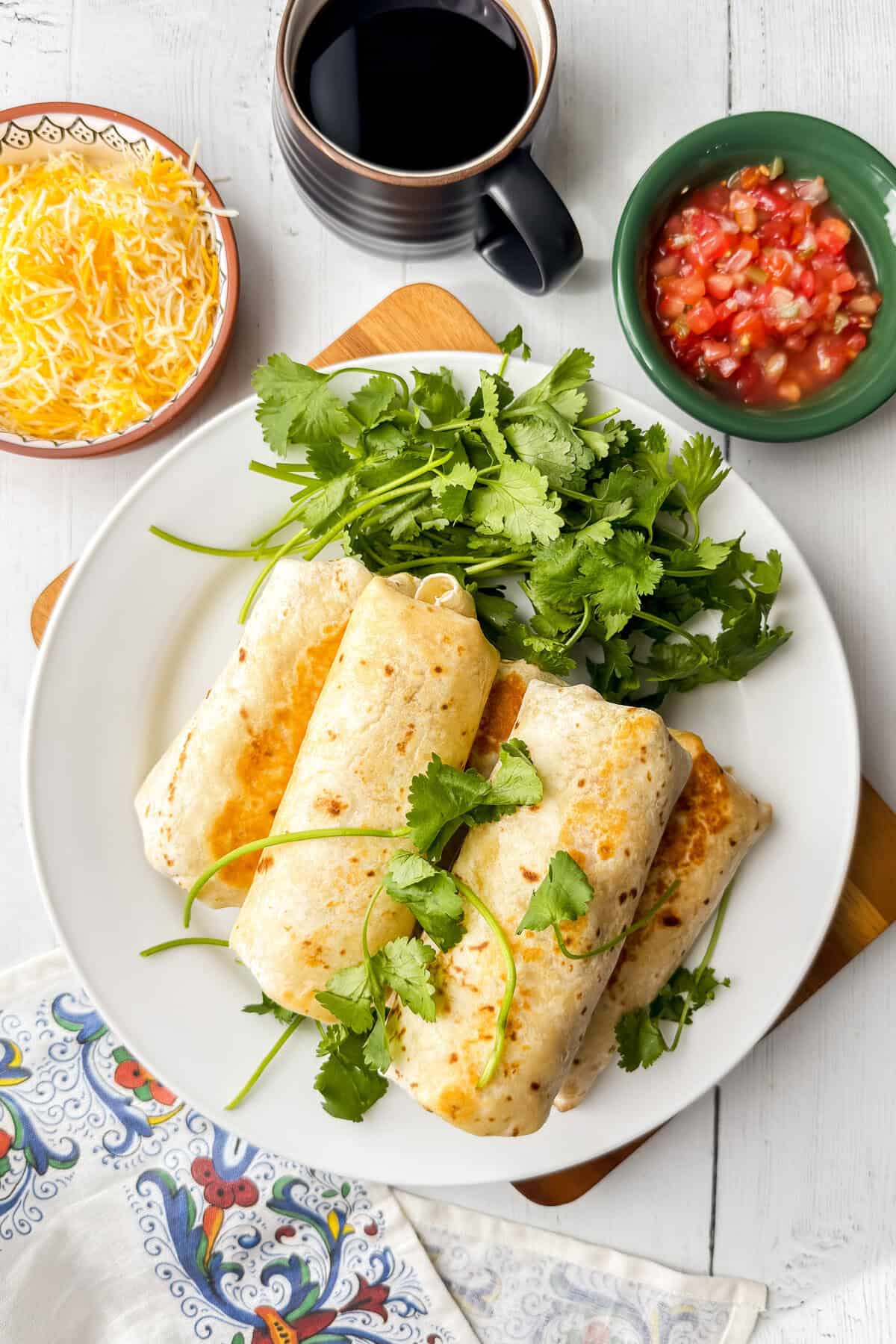 Make-ahead breakfast burritos on white plate with fresh cilantro, shredded cheese, salsa, and coffee.