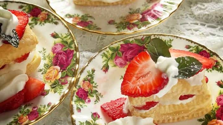 Three floral-patterned plates hold Strawberry Napoleon pastry stacks with cream and strawberries, garnished with mint leaves. The desserts rest on a lace cloth, creating a vintage and elegant presentation.