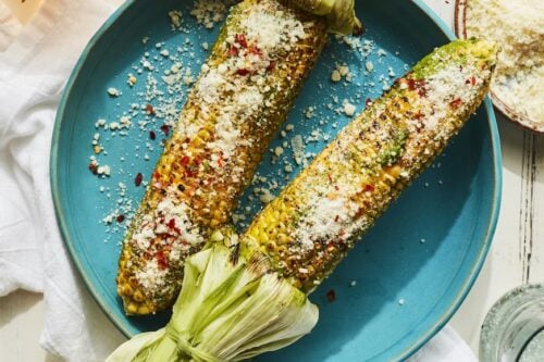Two ears of grilled corn topped with grated cheese and red pepper flakes are served on a blue plate with drinks and condiments nearby—perfect for summer side dishes.