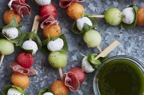 Skewers with watermelon, cantaloupe, honeydew, mozzarella balls, basil, and prosciutto are arranged on a metal surface next to a small bowl of green dipping sauce—perfect for refreshing Summer Side Dishes.
