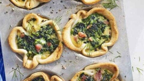 A perfect Mother's Day breakfast: a muffin tray with six puff pastry cups filled with spinach, cheese, and bacon. Fresh dill is sprinkled on top. A blue cloth is visible underneath the tray, and a refreshing glass of orange juice waits in the background.