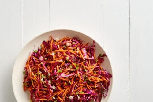 A white bowl filled with colorful shredded carrot and red cabbage salad, perfect for Summer Side Dishes, sits on a white wooden surface with two metal forks resting in the bowl.
