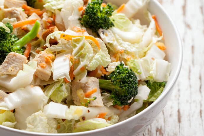 A close-up of a salad in a white bowl featuring pieces of grilled chicken, chopped lettuce, broccoli florets, shredded carrots, and crisp Asian Coleslaw on a rustic white wooden surface.