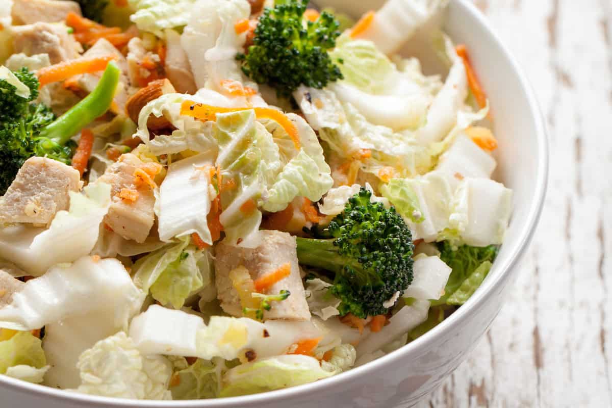 A close-up of a salad in a white bowl featuring pieces of grilled chicken, chopped lettuce, broccoli florets, shredded carrots, and crisp Asian Coleslaw on a rustic white wooden surface.