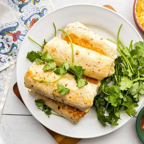 A plate with four Breakfast Burritos garnished with fresh cilantro, next to bowls of shredded cheese, salsa, and a cup of black coffee, on a white wooden table.