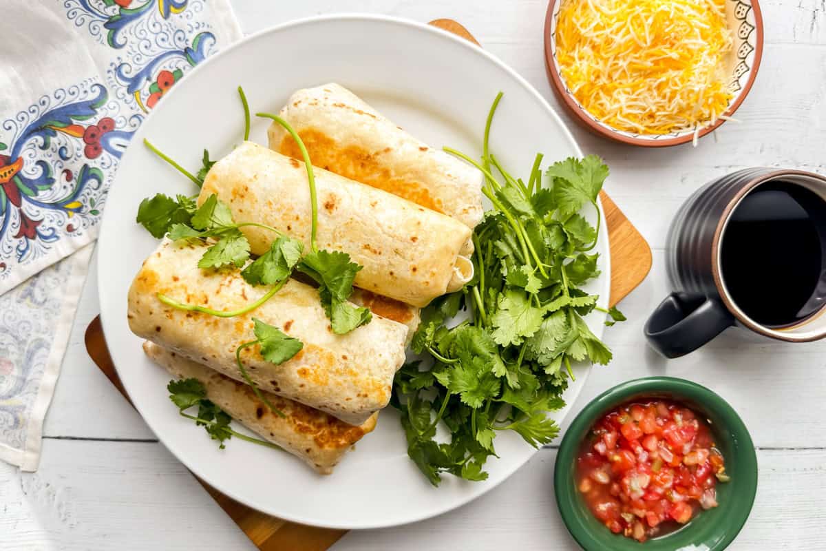 A plate with four Breakfast Burritos garnished with fresh cilantro, next to bowls of shredded cheese, salsa, and a cup of black coffee, on a white wooden table.