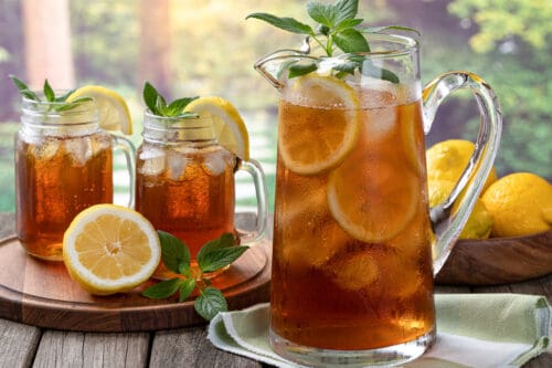 A glass pitcher and two mason jars filled with sweet tea, lemon slices, ice cubes, and mint leaves sit on a wooden surface. Whole and sliced lemons surround the drinks, with a green, sunlit background outdoors.