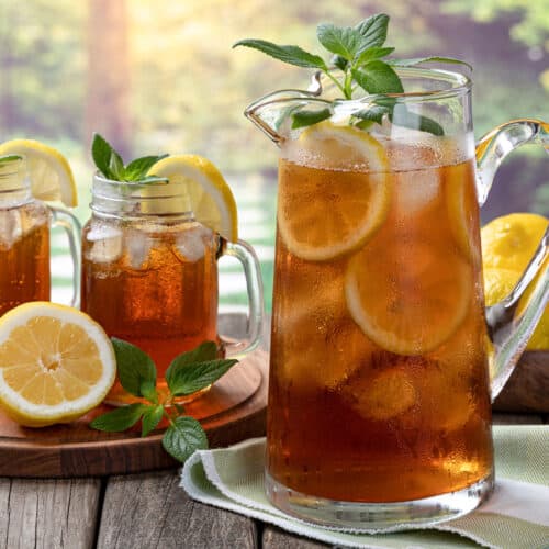 A glass pitcher and two mason jars filled with sweet tea, lemon slices, ice cubes, and mint leaves sit on a wooden surface. Whole and sliced lemons surround the drinks, with a green, sunlit background outdoors.