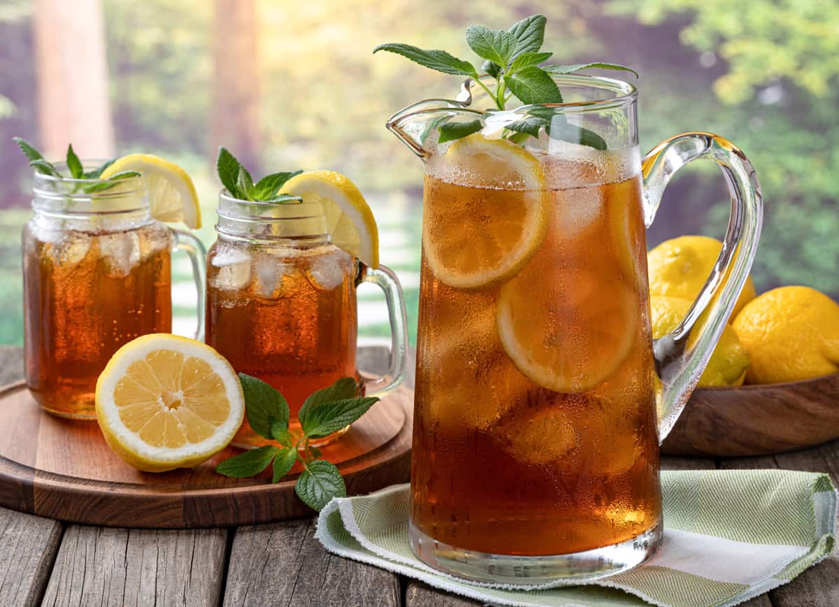 A glass pitcher and two mason jars filled with sweet tea, lemon slices, ice cubes, and mint leaves sit on a wooden surface. Whole and sliced lemons surround the drinks, with a green, sunlit background outdoors.