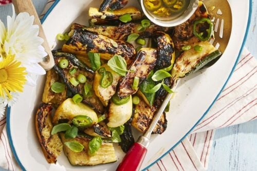 Grilled zucchini slices garnished with herbs, served on a white platter with a small cup of dressing; a red-handled utensil rests on the plate—perfect for light Summer Side Dishes.