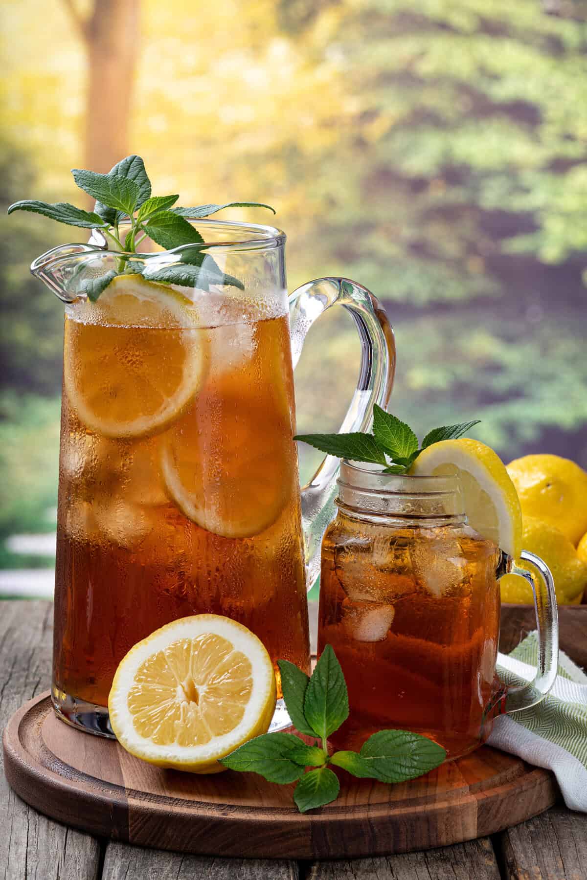 A glass pitcher and jar filled with sweet tea, lemon slices, ice cubes, and mint leaves sit on a wooden board. A halved lemon and mint garnish are nearby, with a blurred leafy background bathed in sunlight.