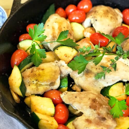 A cast iron skillet filled with baked Chicken Thighs with Tomatoes and Zucchini, featuring zucchini slices and cherry tomatoes, garnished with fresh parsley.