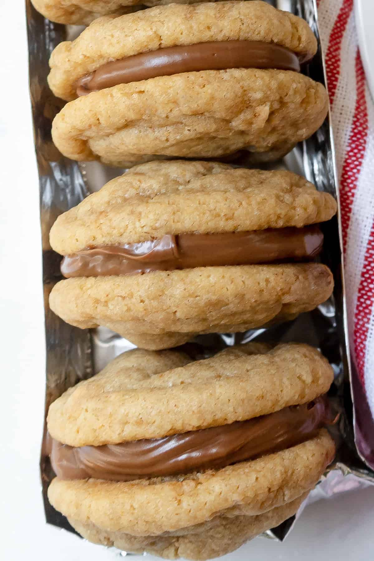 Three graham cookies with light brown, soft-looking texture and a thick layer of chocolate filling are arranged in a row on a tray. A red and white striped cloth is partially visible on the right side.