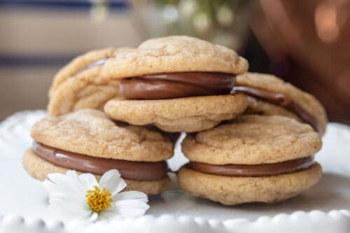 A stack of graham cookies with caramel filling is displayed on a white cake stand, decorated with a small white flower. Blurred flowers are visible in the background.