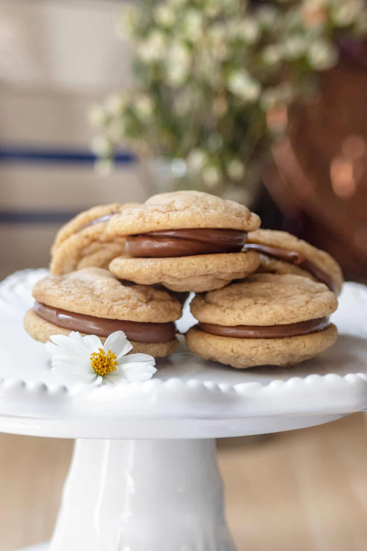 A stack of graham cookies with caramel filling is displayed on a white cake stand, decorated with a small white flower. Blurred flowers are visible in the background.