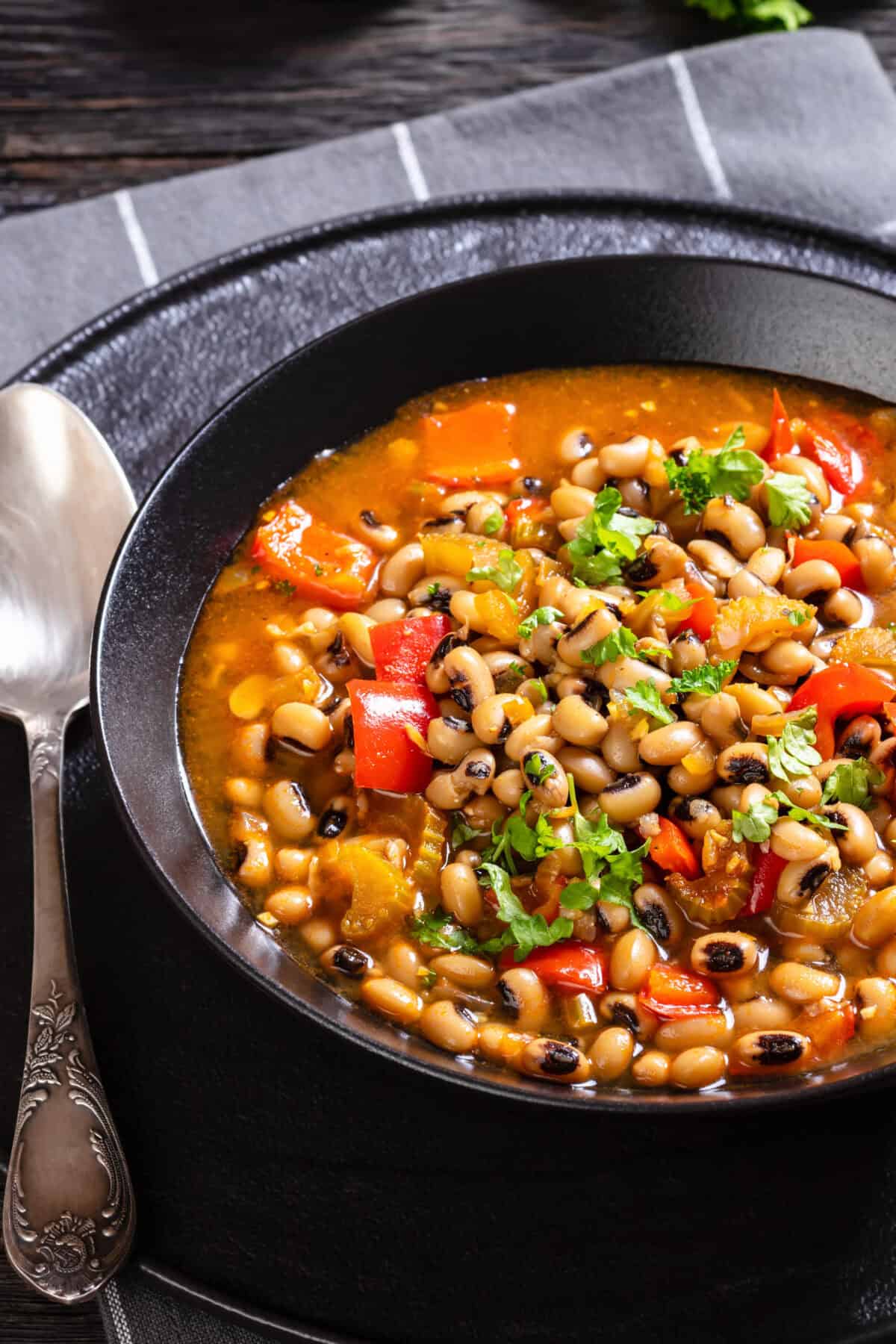 A bowl of Ikarian Stew with black-eyed peas, chopped red peppers, and fresh herbs, served on a dark plate with a vintage spoon beside it on a striped napkin.