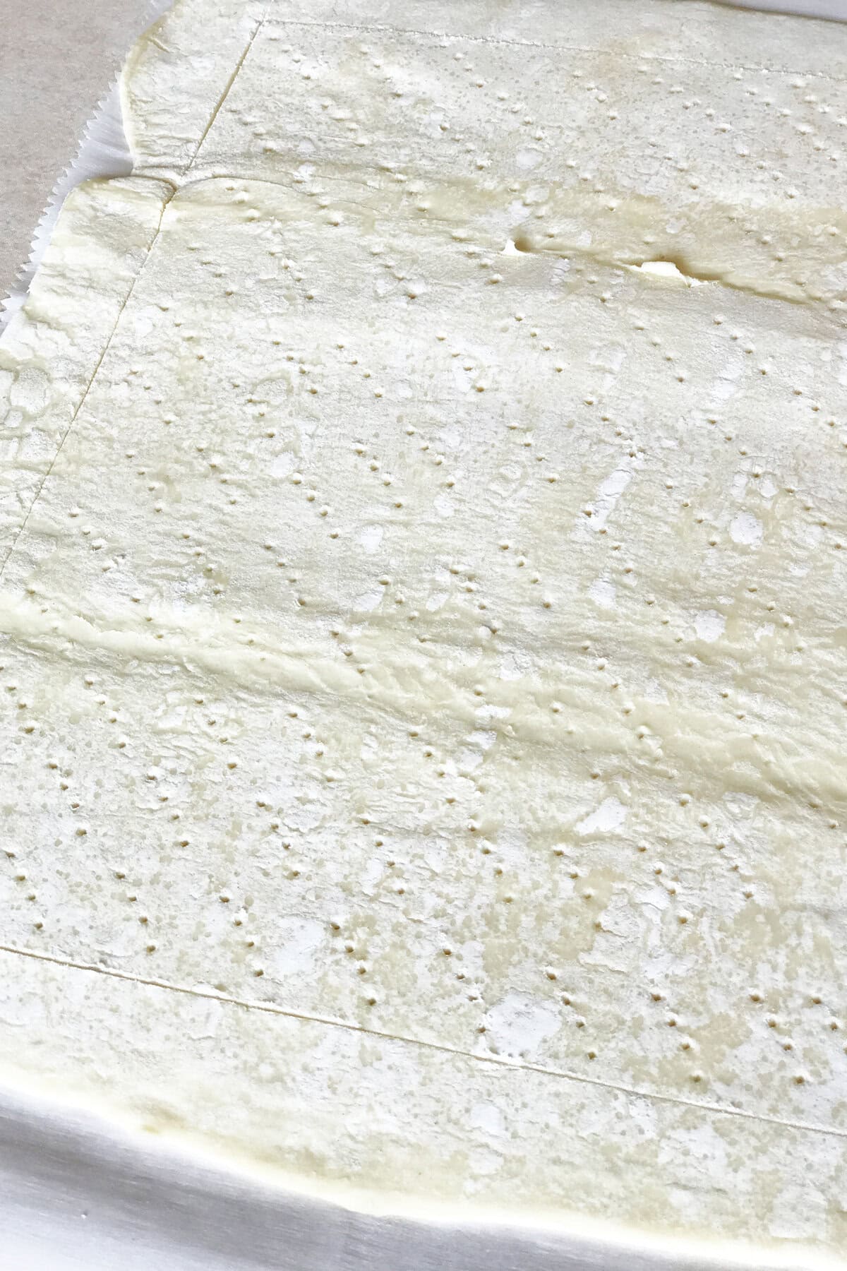 A sheet of raw puff pastry dough on a flat surface, lightly dusted with flour, docked with small holes, and scored into sections with a knife—perfect as the base for a delightful Puff Pastry Strawberry Tart.