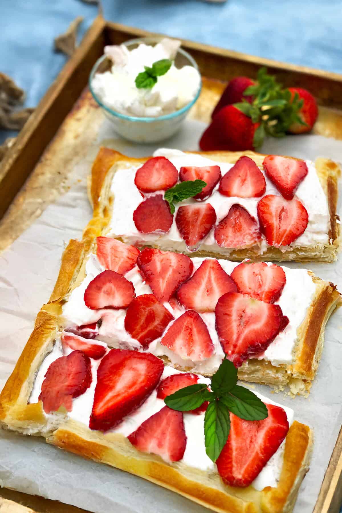 Rectangular Puff Pastry Strawberry Tart slices topped with whipped cream and fresh strawberries are arranged on a tray, one garnished with a mint sprig. A small bowl of whipped cream and whole strawberries sits nearby.