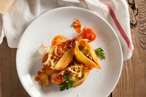 Top view of a white plate featuring stuffed pasta shells with ratatouille sauce, garnished with herbs. A serving dish nearby holds more of this delightful creation alongside a cheese wedge and red-striped cloth. Text overlay reads "Stuffed Pasta Shells & Ratatouille Sauce" with a website link.
