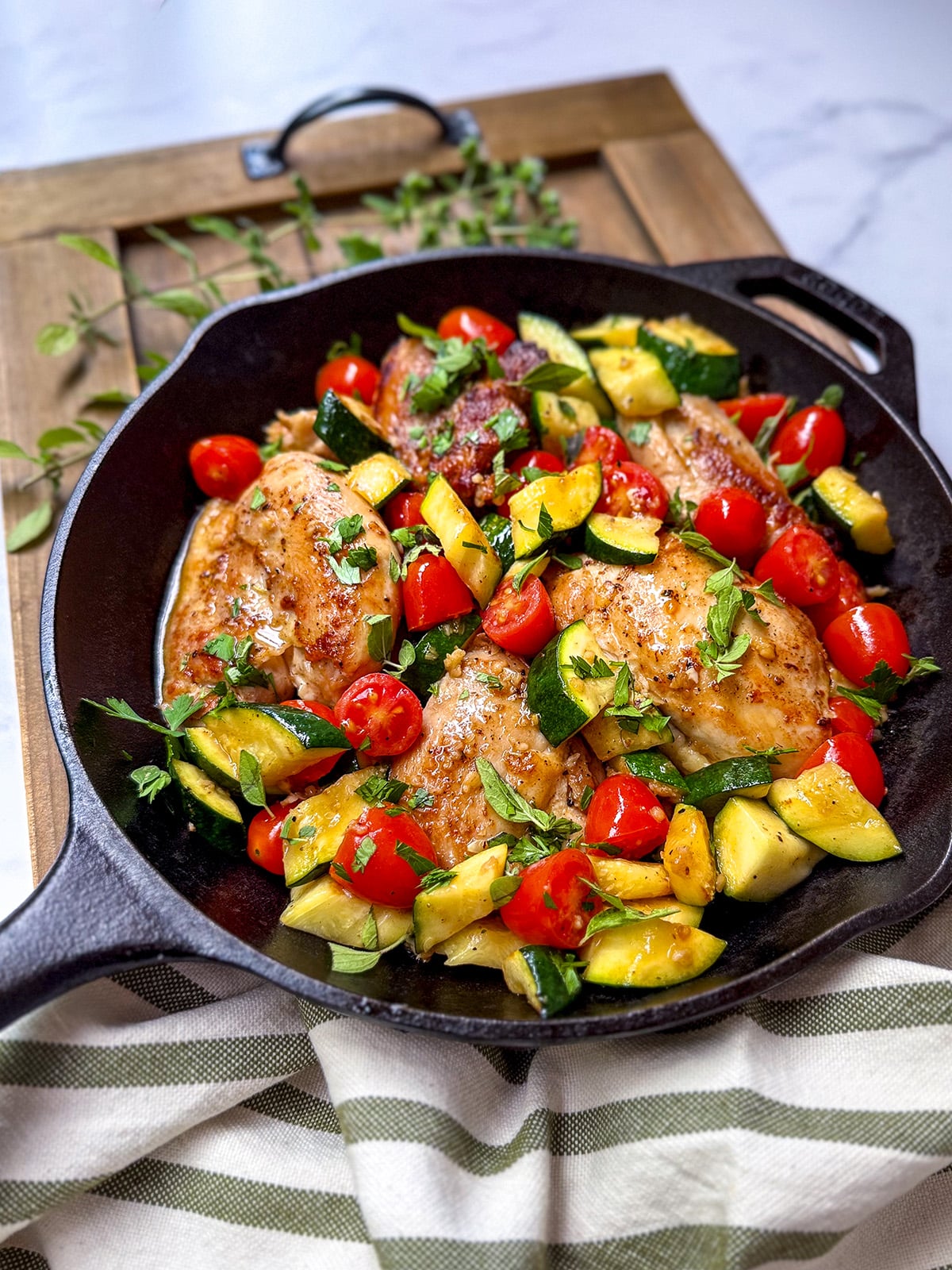 A cast iron skillet with Chicken Thighs with Tomatoes and Zucchini, featuring juicy chicken breasts, set on a wooden board with a striped cloth underneath.