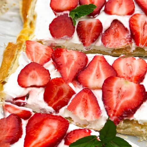 Close-up of a Puff Pastry Strawberry Tart topped with whipped cream and sliced fresh strawberries, garnished with a sprig of mint, sitting on parchment paper.