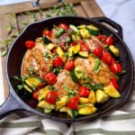A cast iron skillet filled with cooked chicken breasts, chopped zucchini, cherry tomatoes, and herbs on a wooden board creates a vibrant scene reminiscent of classic Chicken Thighs with Tomatoes and Zucchini recipes.