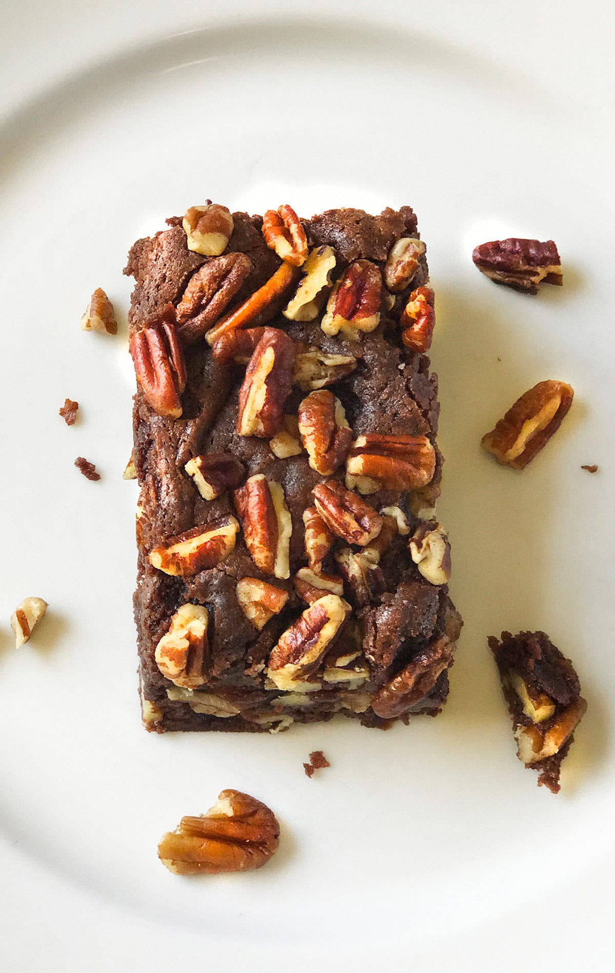 An Espresso Chocolate Brownie topped with pecan pieces sits on a white plate, with a few loose pecans scattered around it.