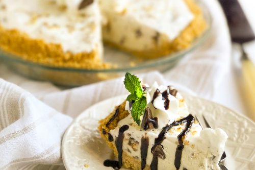 A slice of creamy ice cream pie garnished with mint and chocolate drizzle sits on a white plate, with a fork beside it. The whole pie is in the background on a striped cloth, adorned similarly with whipped cream and mint.