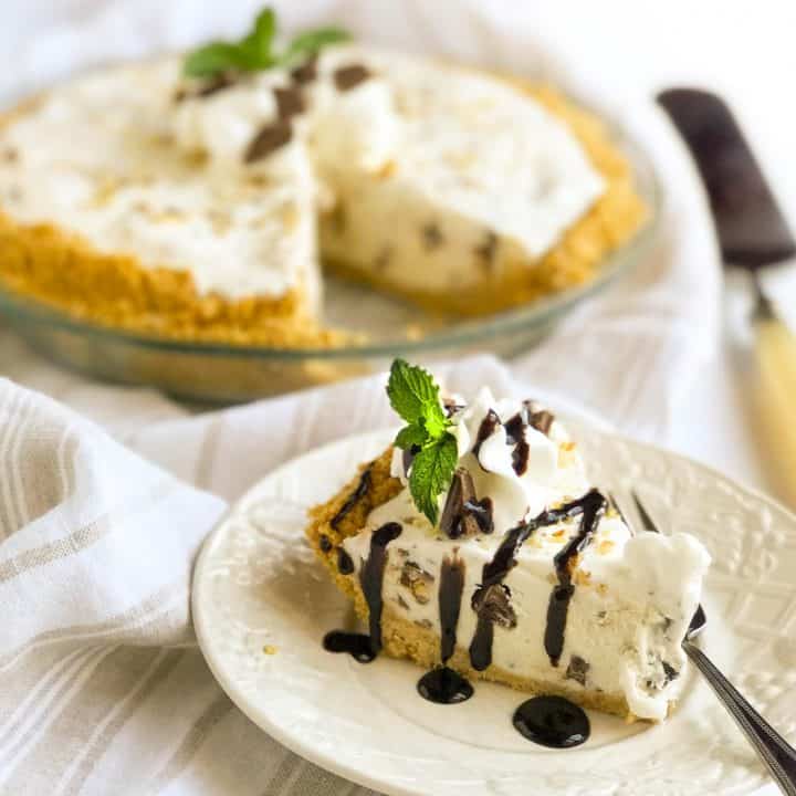 A slice of creamy ice cream pie garnished with mint and chocolate drizzle sits on a white plate, with a fork beside it. The whole pie is in the background on a striped cloth, adorned similarly with whipped cream and mint.