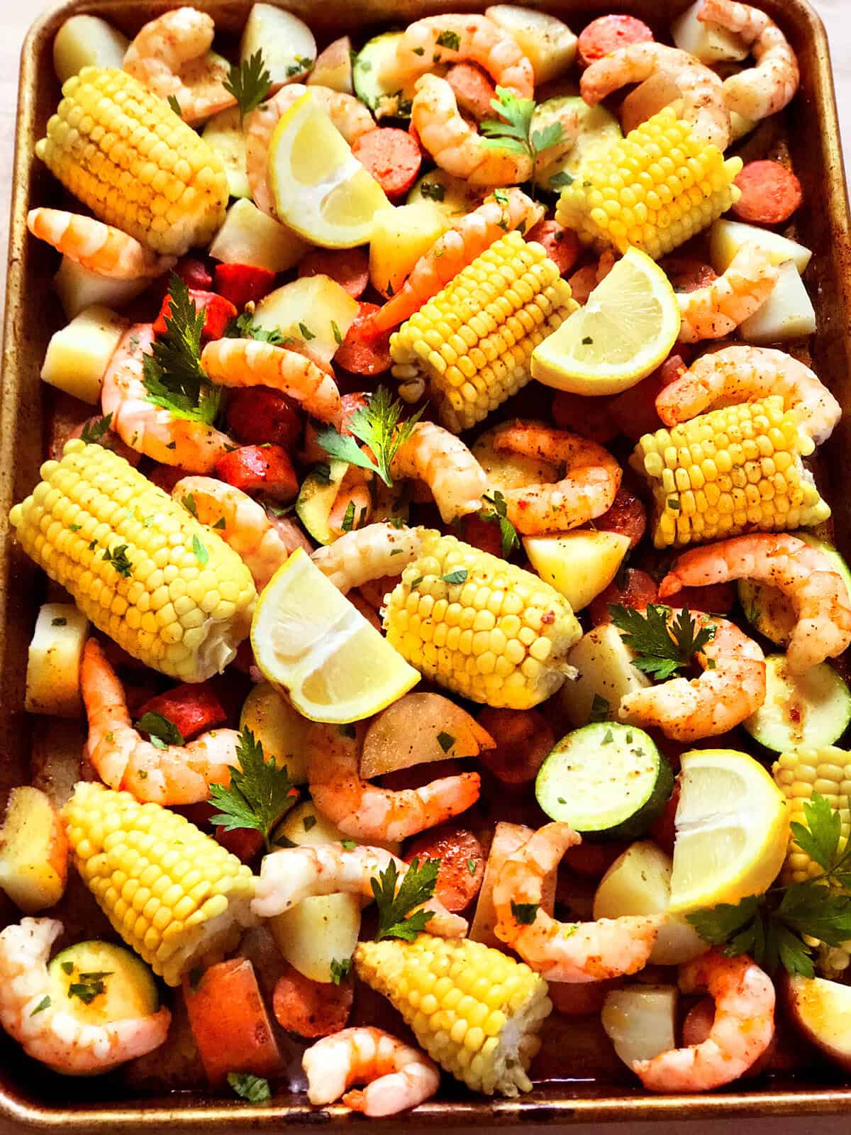 A Sheet Pan Shrimp Boil features cooked shrimp, corn on the cob pieces, potato chunks, sausage slices, lemon wedges, parsley, and zucchini—all mixed together in a colorful, seasoned display.