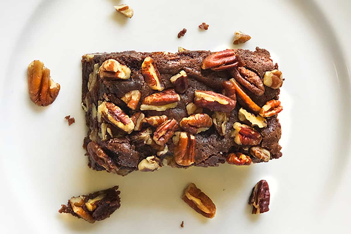 An Espresso Chocolate Brownie topped with chopped pecans sits on a white plate, with a few pecan pieces scattered around it.