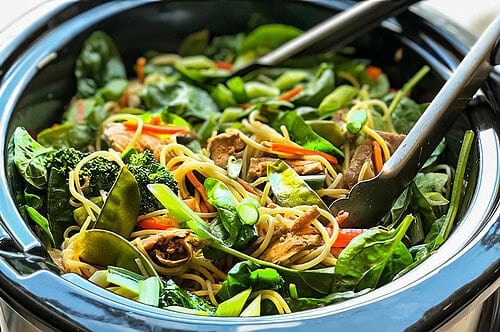 A close-up of a bowl filled with a vibrant stir-fry reminiscent of Slow Cooker Pork Lo Mein. The dish contains noodles, sliced green onions, spinach, broccoli, sliced carrots, snow peas, and tofu. Tongs rest on the bowl's edge, ready to serve.