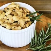 A white ramekin filled with rosemary spiced cashews, garnished with sprigs of fresh rosemary, sits on a wooden board. A bunch of rosemary lies next to the ramekin.